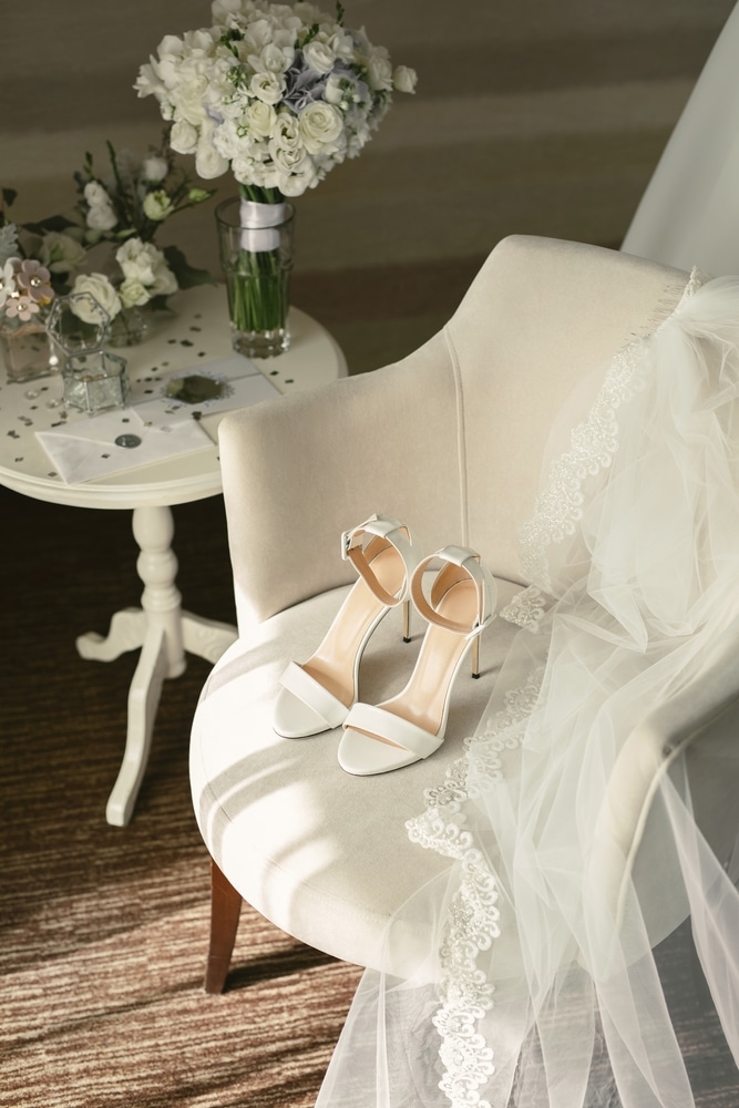 Élégantes chaussures de mariage blanches et voile de dentelle sur une chaise beige. Bouquet de fleurs fraîches et lettre scellée sur la table.